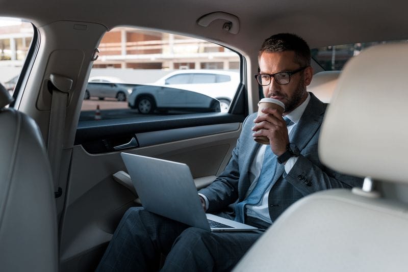 A man in a suit sits in the backseat of an airport transfer car, working on a laptop and drinking from a paper cup.