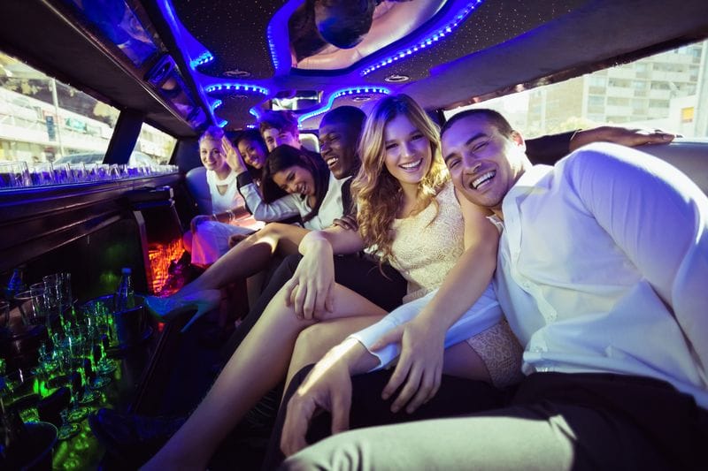 A group of young adults dressed in formal attire sit closely together, smiling and laughing inside a limousine with colorful interior lighting, enjoying their stylish airport transfer.