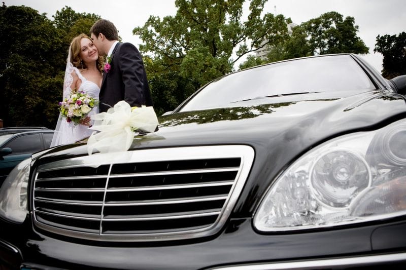 Wedding Limousine 1 A bride and groom stand close together holding a bouquet next to a black car decorated with a white ribbon, as if ready to start their sightseeing tour, with trees in the background.