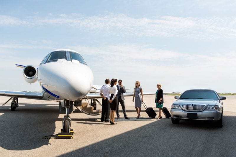 A group of people in business attire stand near a private jet and a parked luxury sedan on an airport tarmac, ready for their exclusive airport transfer.